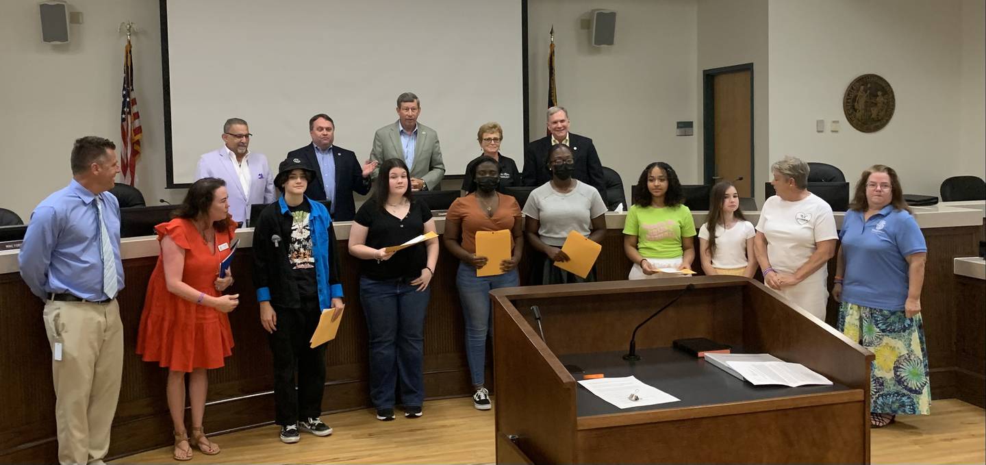 Six children and four staff members stand in front of the long county commissioners table. Five adults in formal clothes stand behind the table. All of them are posed for the photo.